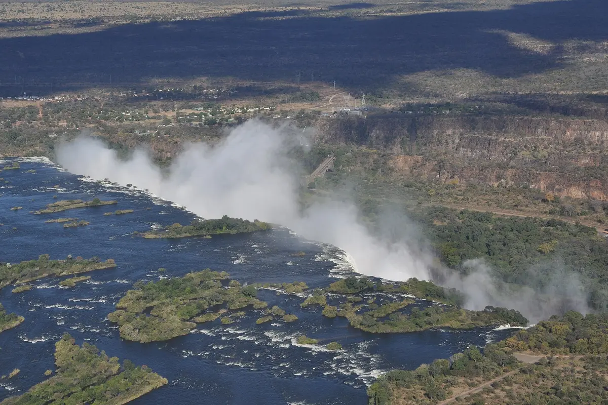 Aerial view of Victoria Falls with spray rising along the line of the falls and the Zambezi above it dotted with tree-covered islands